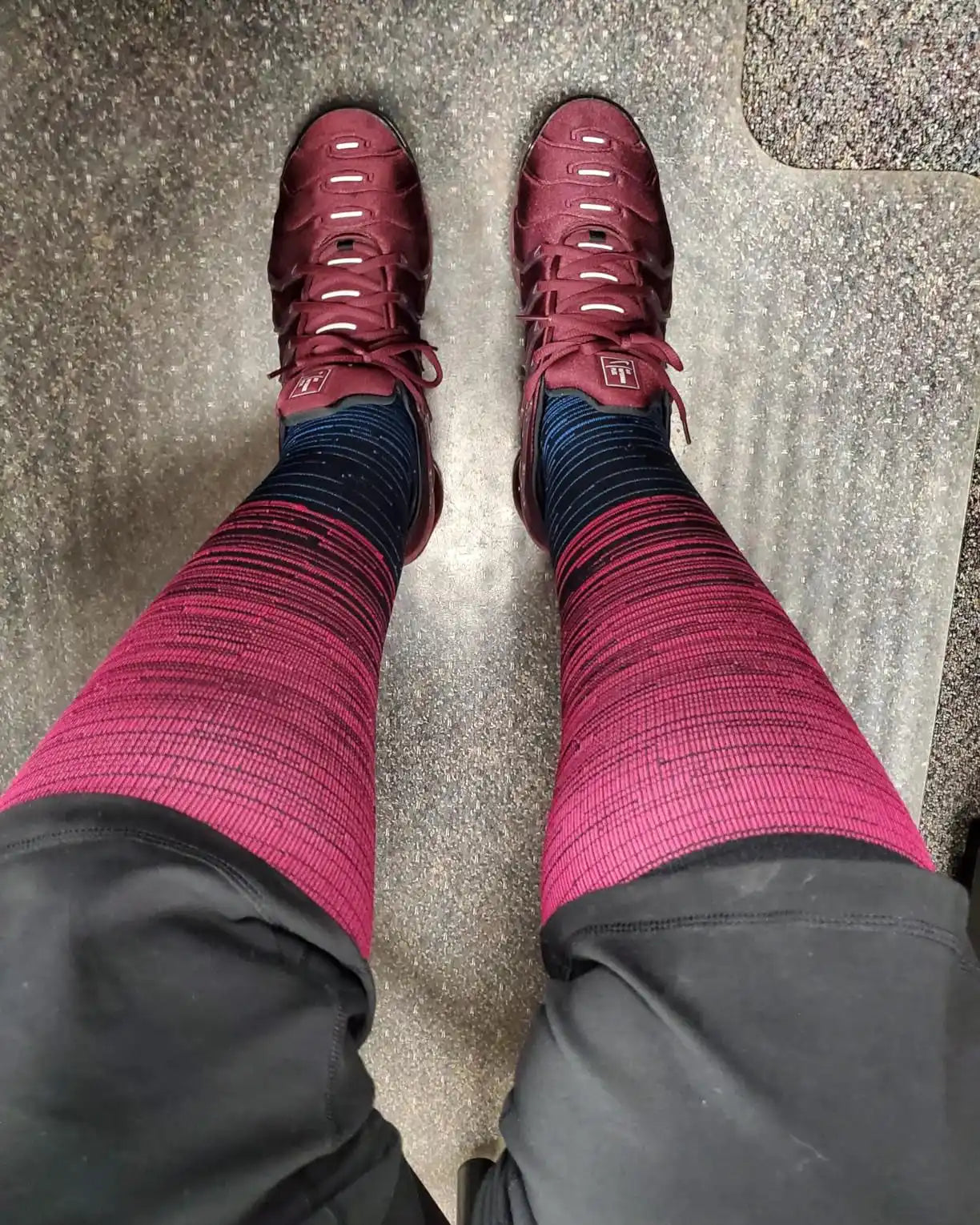 Jambes de la personne en chaussures de sport marron foncé, pantalon noir et chaussettes dégradées noir et rose.